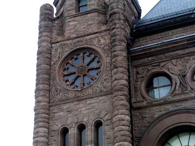 [The stone building has round stone on the corners of a section at the end of the building. That section also has a round window with stone decorations on it. The stone around the window also appears to have a design chiseled in it.]